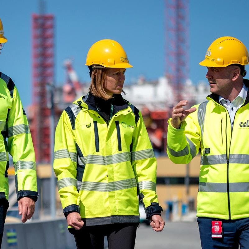 Fire personer i gul vernejakke og hjelm inne på et industriområde med en plattform i bakgrunnen.